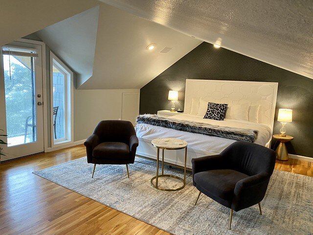 King bed with white bedspread and white headboard. A leopard patterned, square shaped pillow is centered in front of 4 white pillows on the king bed. Two blue chairs sit at the base of the king bed. 