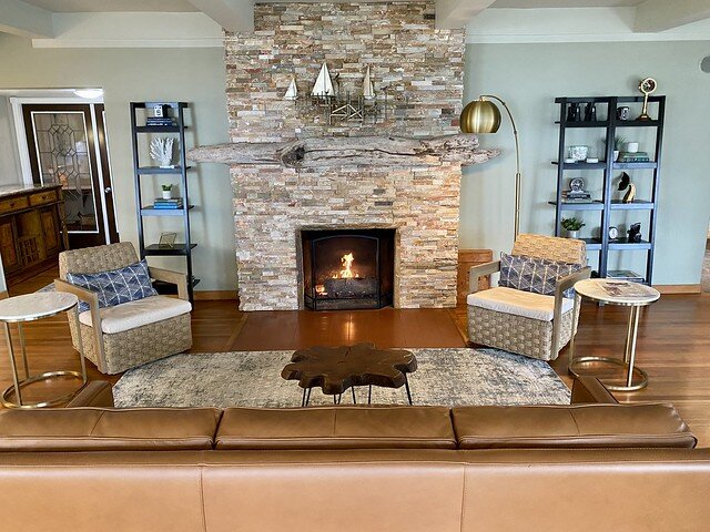 Ocean House living room with hardwood flooring. An area rug is in front of the gas burning fireplace. A light-colored stone wall surrounds the fireplace, rising up from the floor to ceiling. Two wicker chairs flank the gas fireplace. A small, wood slab, dark colored table stands between the fireplace and a caramel colored leather sofa. 