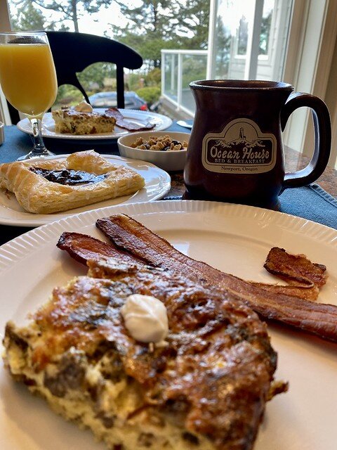 Ocean House mushroom quiche with 2 strips of bacon. A puff pastry square, logo Ocean House coffee mug and glass of orange juice are on the table. An identical meal is across from the food on the same table.