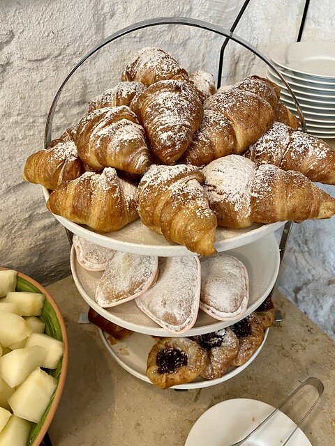 Three-tiered platter of breakfast pastries. Top to bottom; mini croissants with powdered sugar on top, custard tartlet with powdered sugar on top, puff pastry with blackberry jam in center. A plate of honeydew melon is to left of pastry platter. 