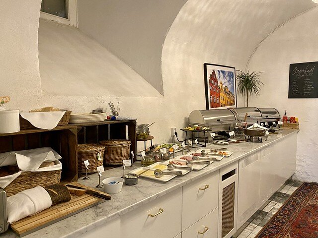 Breakfast buffet with 3 silver chafing dishes, cheese, bread baskets. A photo of Gamla Stan Stockholm colorful buildings hang on the white stucco wall by buffet counter.