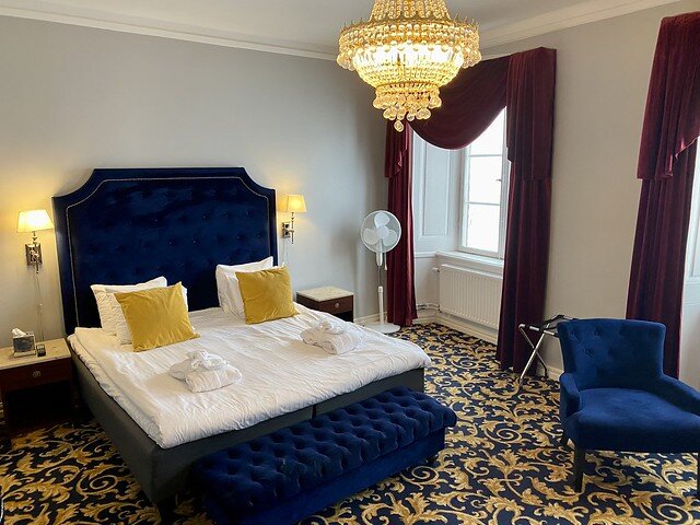 King bed with blue velvet headboard, white duvet, white pillows, yellow accent pillows , 2 white robes on bed. Blue and gold carpet. Burgundy velvet drapes on 2 bedroom windows on right side of room. Glass, tear drop chandelier in center of bedroom. Small, blue velvet chair to right of bed, by window. Floor stand fan by back window, right of bed. 