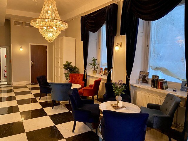 Hotel lobby with chandelier, two round, white marble table tops. Six blue velvet chairs surround two tables. Black and white checked marble tile on floor. Two picture windows framed by dark, velvet drapes. 