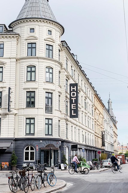 Five-story Ibsens Hotel exterior. White building with 8 rectangular windows. The ground floor of the hotel is painted gray with a front entrance. A rack of parked bicycles are across the street. Two bicyclists pass in front of the hotel.