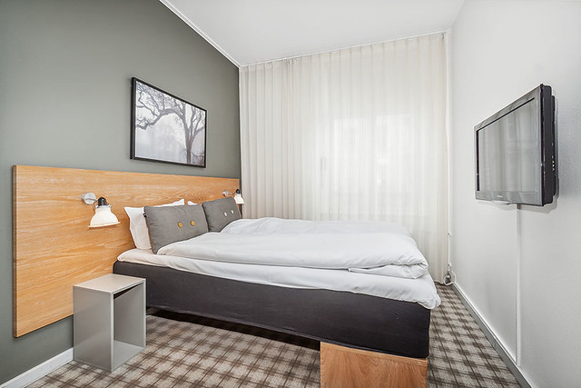 Small hotel room with semi-double bed. Gray wall, light wood headboard. White duvet with 2 white pillows and 2 gray throw pillows. A beige carpet with brown pattern is under the bed and flat screen tv, mounted on the wall, across from the bed. 