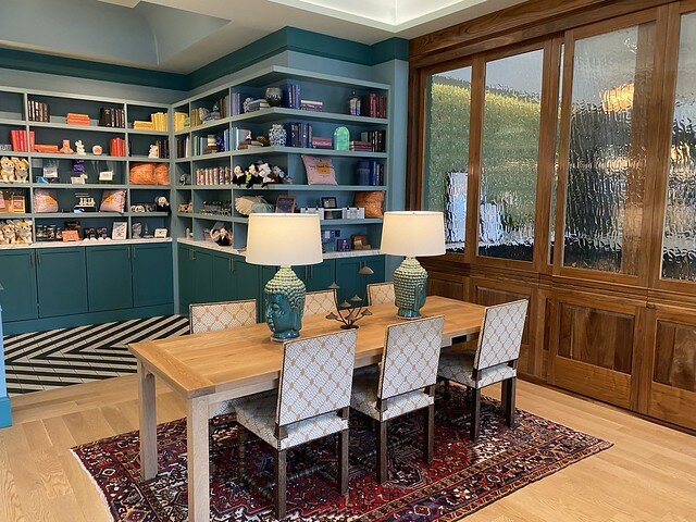 Gift shop looks like a dining room in hotel lobby. Wood dining room table has 6 chairs, 3 on either side of table. Two blue lamps are on dining room table. Trinkets and books are displayed on 5 book shelves. A wood and glass sliding door separates gift shop from hotel restaurant. 