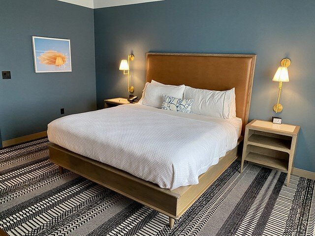 King bed, white duvet, white pillows. Leather headboard. Blue walls, black and white patterned area rug underneath bed.