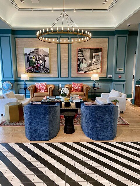 Black and white floor tiles connect to oak flooring in hotel lobby. Two pieces of modern art are on the wall. Two blue chairs are across from two light brown, leather chairs, with pink throw pillows, in lobby. Circular light fixture hangs from ceiling. 