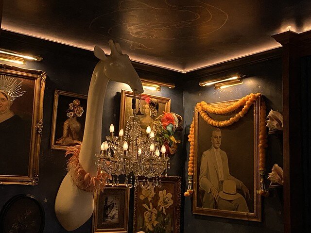 Porcelain giraffe head is surrounded by 6 framed pictures of men and women on the wall. An ornate crystal chandelier hangs from chain in giraffe's mouth.