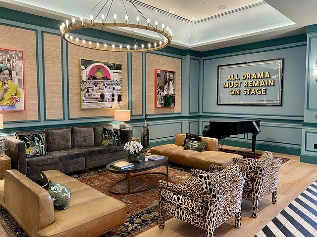 All Drama Must Remain on Stage artwork hangs next to baby grand piano in hotel lobby. Two leopard-skin chairs are next to 2 beige leather sofas. Drake brown fabric sofa with 2 floral throw pillows across from leopard upholstered chairs and oval coffee table. Circular light fixture above coffee table. Three pieces of modern art hang on the back wall. 