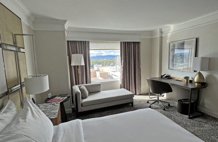 White hotel bed in a beige room with floor-to-ceiling window showing distant snow-covered mountains