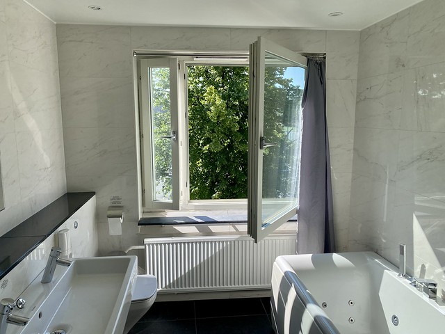 White marble bathroom walls surround large, open picture window at Hotel Allen. Left of window is white ceramic farm-style double sink. To the right of open window is white jetted bathtub. 