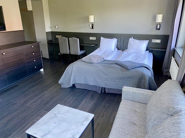 Family room with two single beds, partial views of gray tweed sofa bed and rectangular metal table with white marble table top. Black, faux wood cabinet with mini fridge to left of beds. 