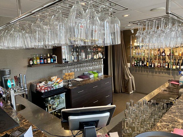 Airport hotel bar with beer glasses, wine glasses and champagne flute glasses hanging upside down from metal rack. More glasses are on the granite bar countertop. The back of a computer screen is centered below the glasses. Across from the counter is a wall with a shelf of alcohol bottles, and more glasses. To the right is a three-tiered glass shelf of more alcohol bottles. 