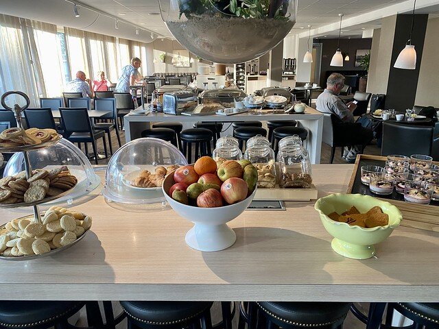 Hotel breakfast buffet with bowl of apples, tray of several individual glass bowls of yogurt with muesli and blueberries on top. A 3-tiered cookie tray is to the left, as well as a plate of croissants, covered in a plastic dome. Beyond is a counter with toaster, fresh bread, hardboiled eggs. Table beyond holds cheese, meats and mini croissants. To the left are tables with 2 black chairs on either side of tables. Three hotel guests are seated at two tables. A man checks his smart phone screen to the right of the buffet table. 