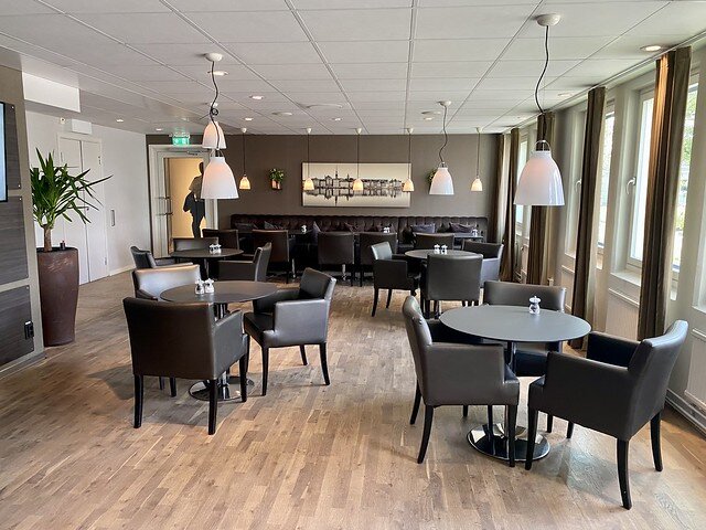 Hotel lobby with random plank hardwood floors. Four small, circular tables, with 3 brown leather chairs at each table, surround the tables. Glass picture windows open, to the right of the room. Four glass, dome, bell-shaped, white light fixtures hang from the ceiling, above each table. A black suede banquette sofa is on the back wall with 3 leather chairs facing the sofa. A hallway on the left, back of the room, leads to hotel rooms. 