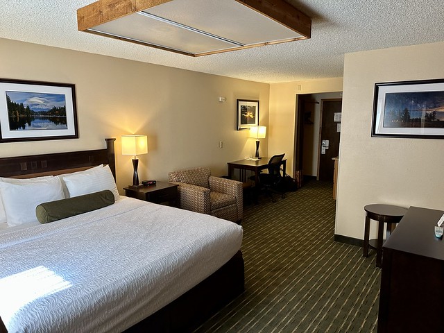 BW Tree House king room with white bedding, small wood nightstand with lamp. Next to nightstand is plaid cloth chair. A black desk and ergonomic black chair are next to plaid chair. Framed art of local Shasta nature scenes hang on the walls. 