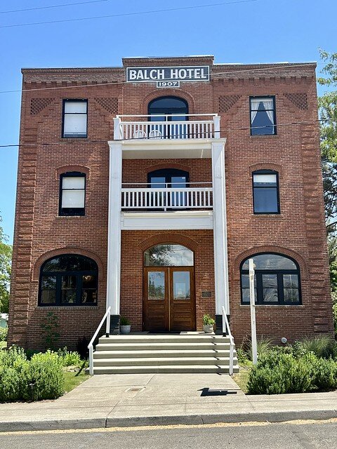 Three-story red brick, hotel with white block letters on the top of the building, reading Balch Hotel 1907. Rectangular shaped glass windows are on either side of the top 2 stories of the hotel. Arcade-shaped windows are in the center of the brick building on top 2 stories. Ground floor glass door front entrance to the hotel is framed with 2 white banisters, serving as the base of the top two balconies over the 2nd and 3rd floor. Several concrete steps lead to the front porch hotel entrance.