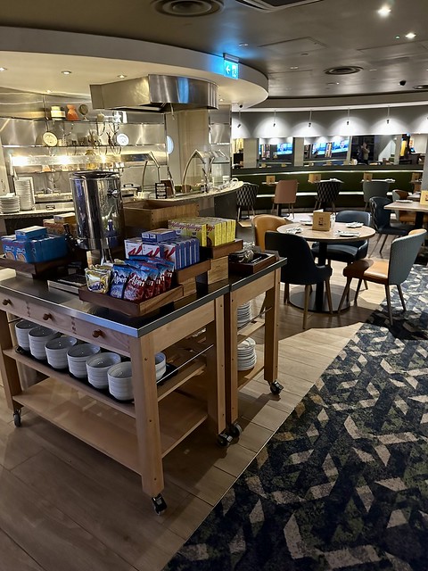 Holiday Inn London breakfast buffet. Cold cereal station with cereal bowls under the wood table. Behind the square table is the stainless steel hot buffet, for omelettes. A round table with 4 chairs is in front on the buffet station. 