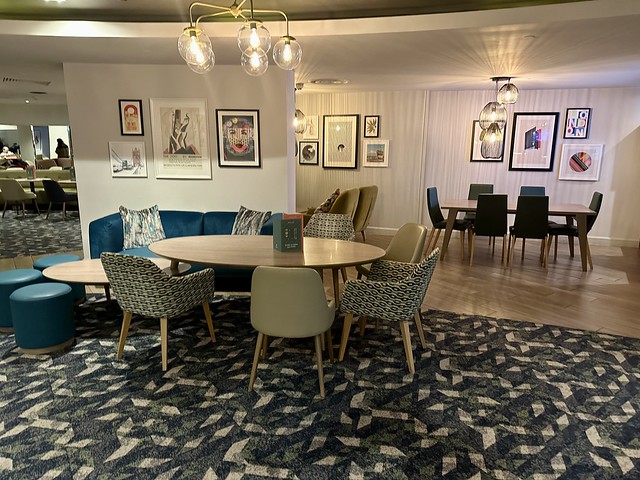 Restaurant and lounge area at Holiday Inn London Regent's Park. An oval wood table with 5 chairs is next to a blue leather sofa banquette and lower round table with 3 blue stools. To the right is another wood dining table with 6 chairs. 