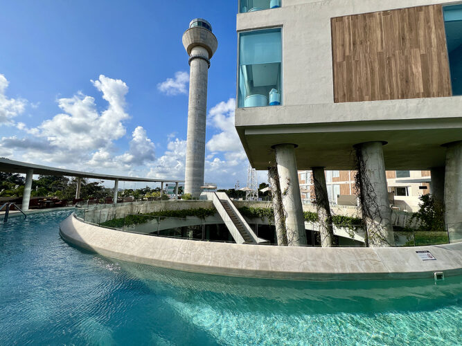 Curved pool underneath hotel rooms with tall tower under blue sky. 