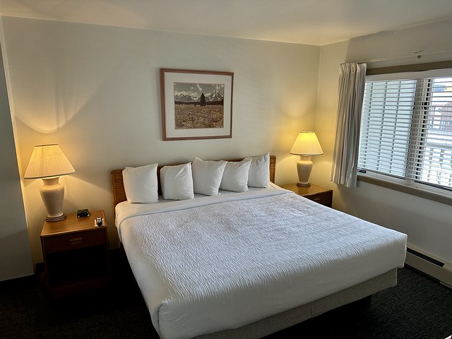 King bed with white bedding and 4 white pillows. A beige desk lamp with white lampshade was on either side of the bed. A color, framed photo of Jackson, Wyoming's Grand Tetons, dusted with snow, hung above the king bed. 