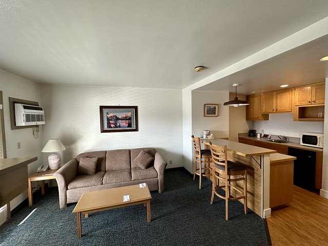 Ranch Inn kitchenette with sink, mini fridge, microwave oven and kitchen counter with 2 chairs. Beyond the kitchenette was a milk chocolate colored fabric sofa bed. An end table with lamp was left of the couch. A light wood coffee table was in front of the couch. Legend of the Lake photo of Jackson, Wyoming lake hangs above the couch. 