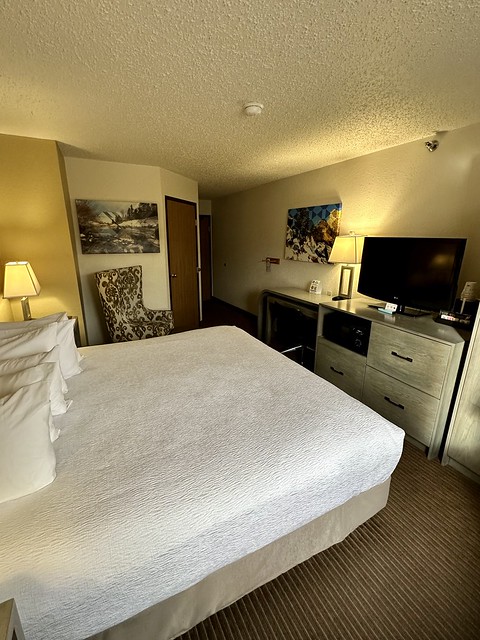 Best Western hotel room with king bed & 5 small white pillows on the white bedspread. A dresser held a small microwave oven and flat screen tv on top of the dresser. A wing-back chair was wedged next to the bed and the bathroom door. 