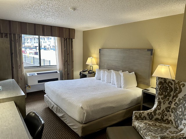 Best Western Alpenglo king room with white bedspread. Six, square white pillows are lined up neatly under the gray wood king headboard. Small, wood bedside tables hold metal lamps with lampshades. A picture window looks out to the parking lot. A room size heater and air-conditioner unit is mounted under the window. 