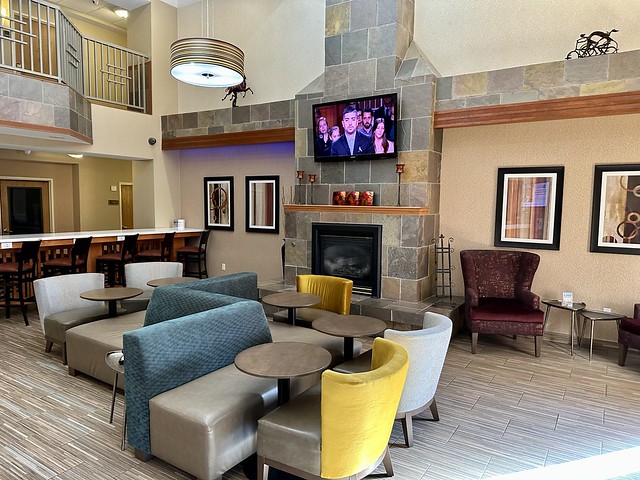 Spacious 2-story hotel lobby with gas fireplace, 5 small, circular tables and a grey and blue banquette and chairs in front of the fireplace. A large, flat-screen tv is bolted to the stone fireplace. 