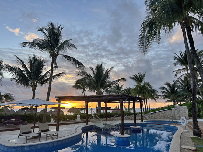 Palm trees around a pool in front of the beach with sunset at Excellence Riviera Cancun