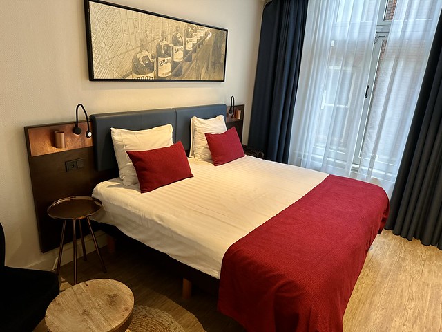 Double bed with white duvet and red runner at base of bed, with 2 accent red pillows. Artwork of former liquor factory bottles above bed. 