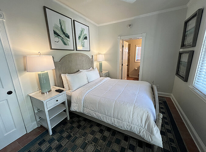 Hotel room with queen bed and white linens in small room with white nightstand.