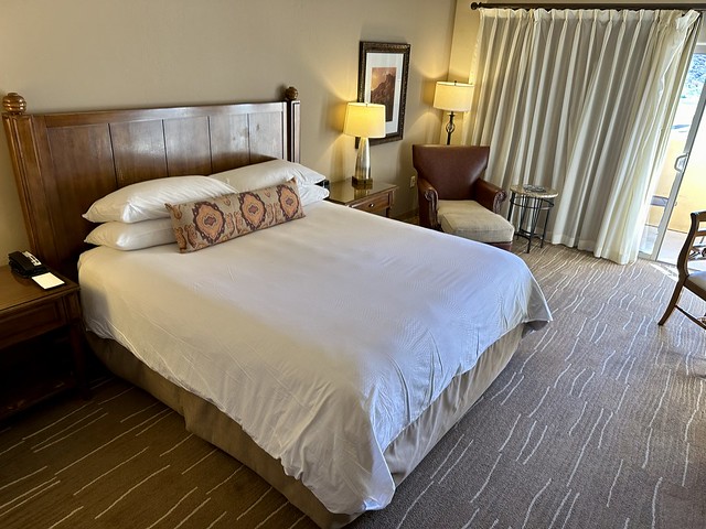 King room features white bedding, southwest decorative pillow, wooden headboard. End table on either side of bed and brown leather chair with beige cloth foot stool. Linen curtains shade the floor-to-ceiling sliding window, opening to the small balcony. 