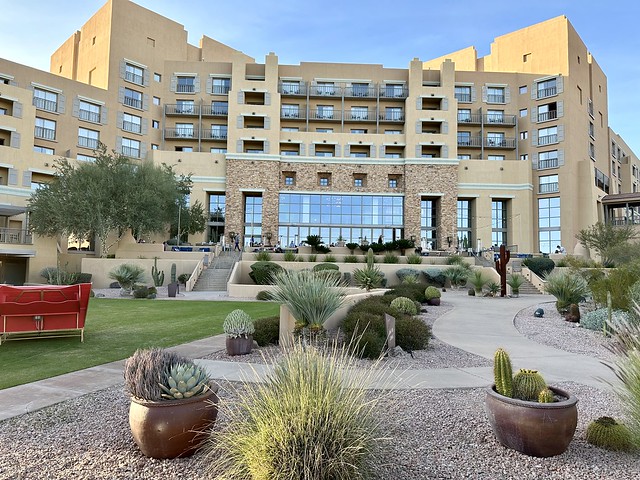 Multi-story JW Marriott Starr Pass Resort with 3-story lobby windows looking out to Sonoran desert. Small cactus punctuate the desert landscape. A large olive tree is to left of open-air terrace. 