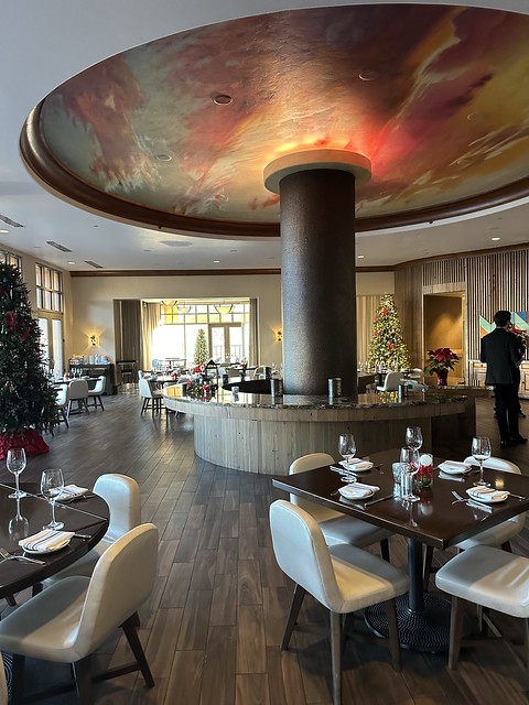 Signature Grill with a center metal pillar in the middle of the restaurant floor. Tables set with place settings and wine glasses for 4 persons are on the dark wood floor. A holiday Christmas tree is behind the steel pillar and to the right. A restaurant employee in a black suit has his back to the camera. 