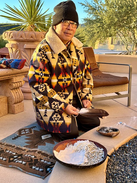 Navajo tribe member Larry Redhouse wears a fleece jacket with Native American tribal patterns of yellow, black, red and beige colors. A black beanie covers his head. He is kneeling on a Navajo blanket and holding an eagle feather in his right hand. A large bowl holds sage, string and small squares of cloth to make our prayer ties. Incense burns in a smaller bowl, under the eagle feather. 