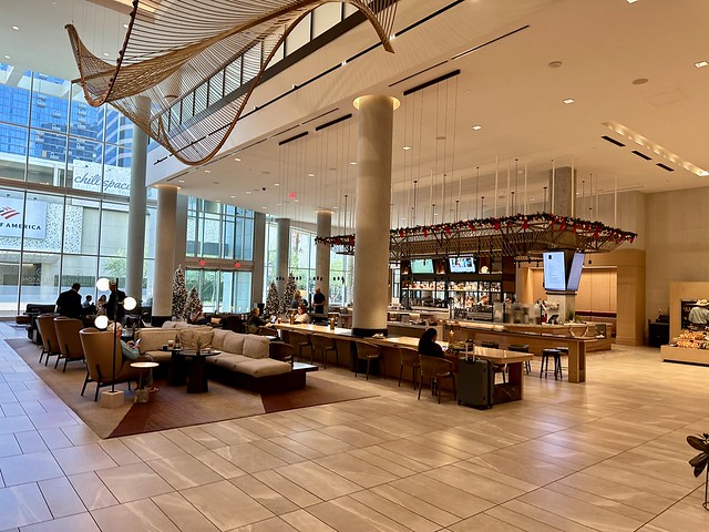 Sheraton Phoenix Downtown hotel lobby with a woman seated next to her suitcase at one end of the wooden community table. A man and woman sit across from each other at the opposite end of the community table. A male hotel guest sits in one of three beige leather chairs across from an l-shaped sofa. Two small, round circular tables are in front of the chair and sofa. 