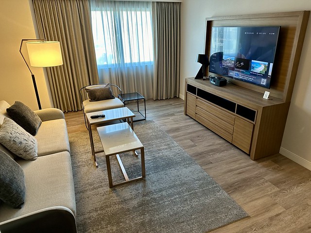 Mountain view king suite with sofa sleeper bed, two small rectangular tables and one modern chair on a area rug. A wooden entertainment center holds a 65 inch flat screen tv. 