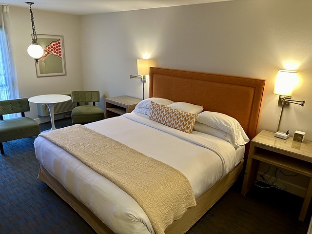 King bed with modern retro-chic design and orange headboard. Modern art piece on wall and white plastic circular table with lime green, velvet chairs on either side. 