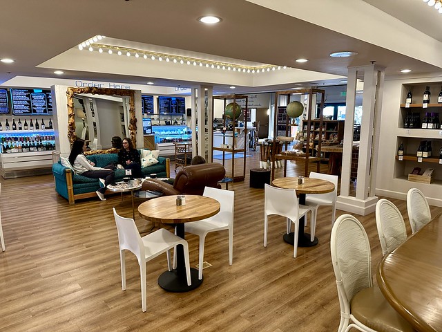 Luna Cafe with wooden, circular tables and a plastic white chair on either side of dining table. Two women drink coffee on a blue velvet sofa in front of the Luna Cafe to-go counter. 