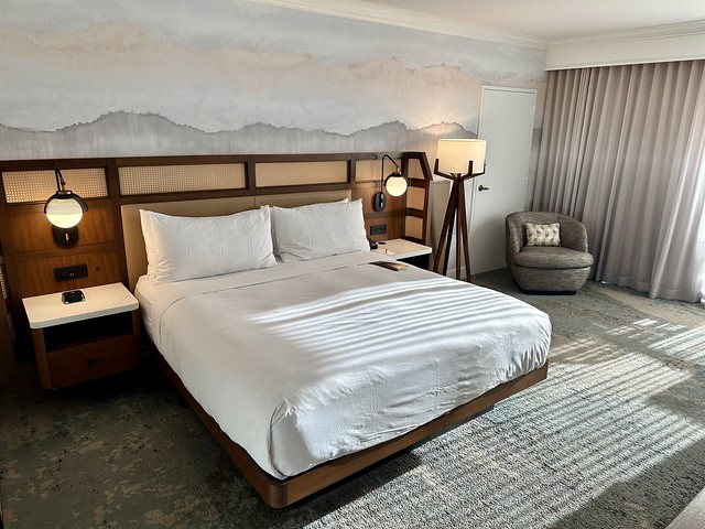 King bed with white bedding, 4 white pillows and wood headboard. A mountain mural is displayed behind the headboard with blue, gray and white colors. A black metal floor lamp is to the right of the bed. 