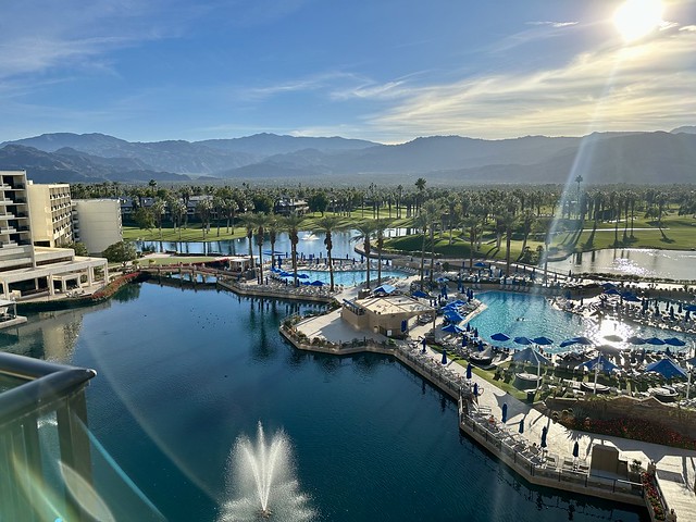 The JW Marriott Desert Springs Resort is partially shown to the left of the photo. A man-made lake has a fountain sending water into the lake. A cement walkway separates the lake from two swimming pools. Beyond, green grass and palm trees stretch to the base of the Santa Rosa Mountains. 