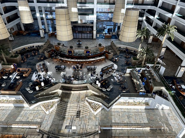 Looking down from the 8th floor of JW Marriott Desert Springs hotel into the lobby. A sushi bar and cocktail lounge are in the spacious open-lobby with tables and chairs placed about the cavernous space. 