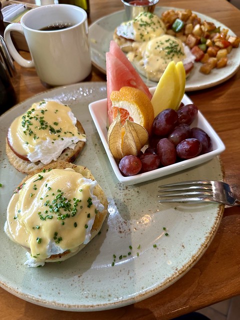 Eggs Benedict English Muffins with Canadian Bacon, poached eggs and hollandaise sauce. Fresh fruit cup of purple grapes golden gooseberry, watermelon slices and mango slices are also on the breakfast plate. 