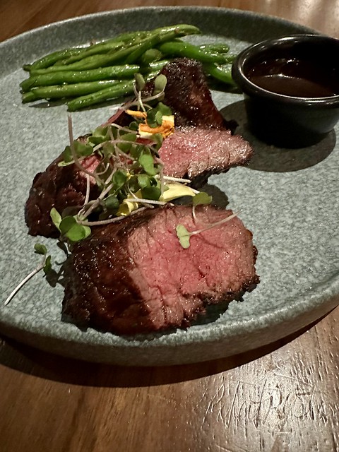 Hanger steak, cooked medium rare, with micro greens, green beans and a sauce in ramekin on ceramic green speckled plate. 