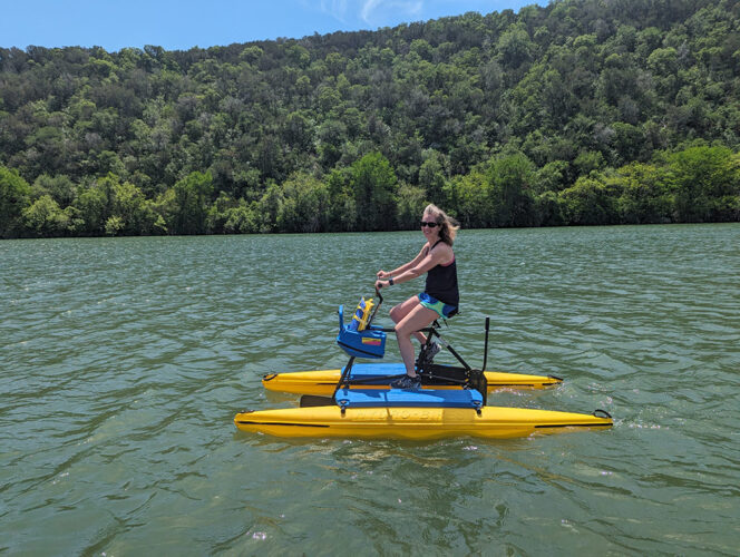 Woman in tank top and shorts on a pontoon bike on a green-blue river with forest.