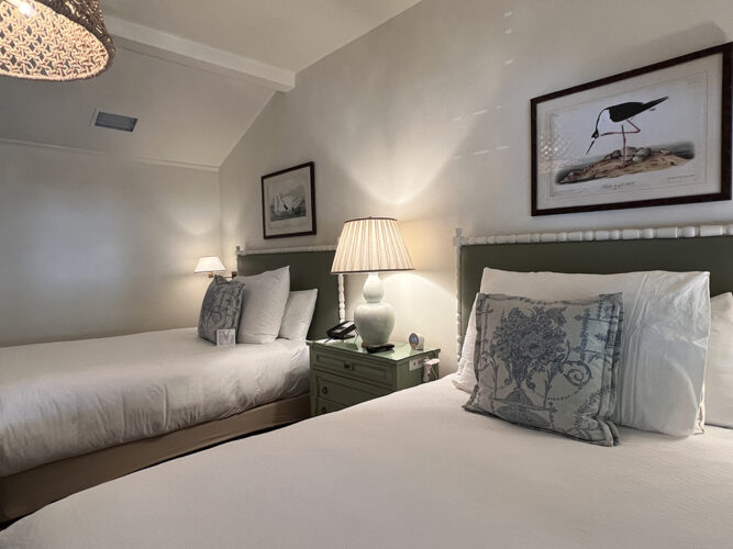 Two queen hotel beds with white comforters in a room with white walls.