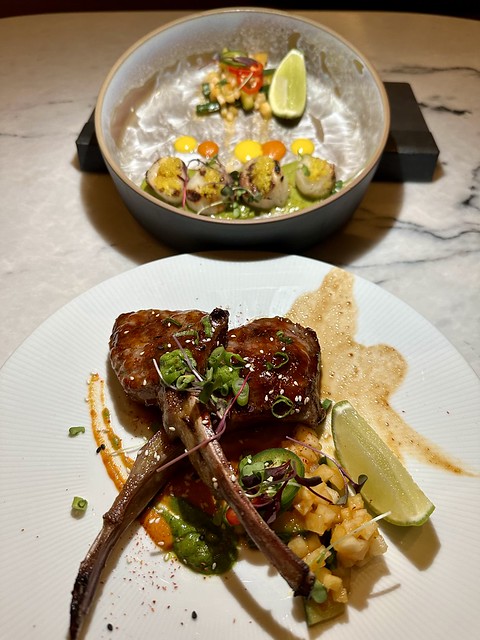 Plate of 2 lamb chops, in addition to a plate of four broiled scallops with orange and yellow dots of sauce that accompany a small mound of mango chutney. 