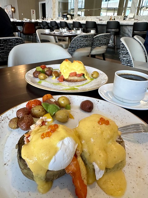 Lobster eggs Benedict on 2 artichoke hearts with Hollandaise sauce drizzled on top of 2 poached eggs. Behind the front plate is a separate plate of traditional Eggs Benedict on 2 English Muffins. A cup of coffe on white saucer is to right of food plate. In the background is a white marble counter at Fountain Restaurant inside Signia by Hilton San Jose. 